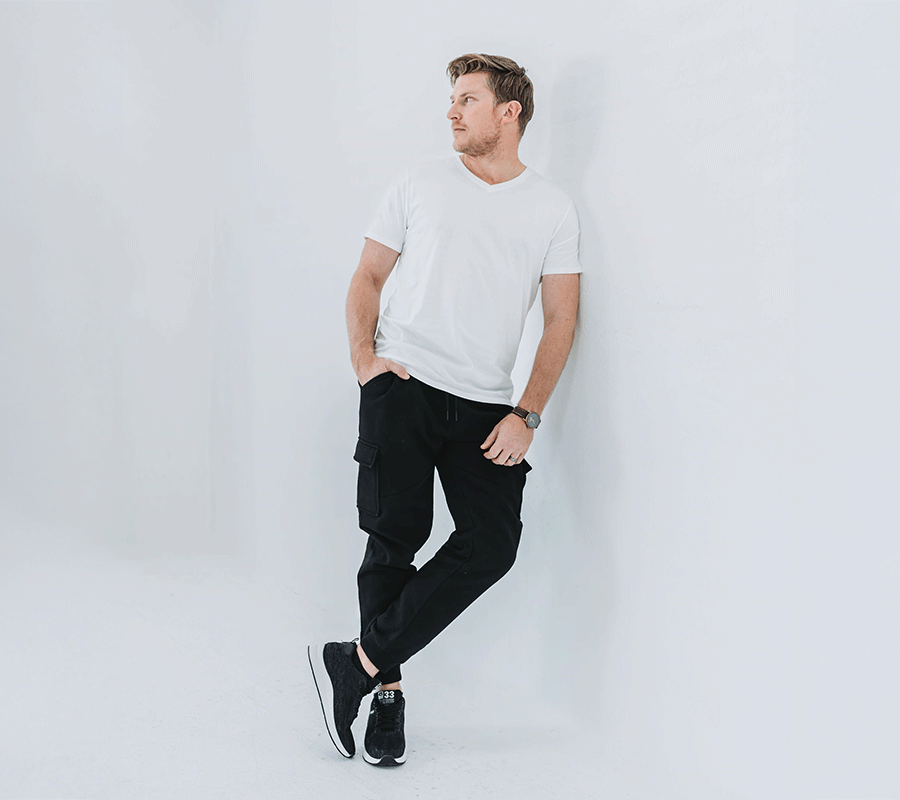 A casually dressed man leans against a light gray wall, wearing a white V-neck t-shirt, black cargo pants, and black sneakers.
