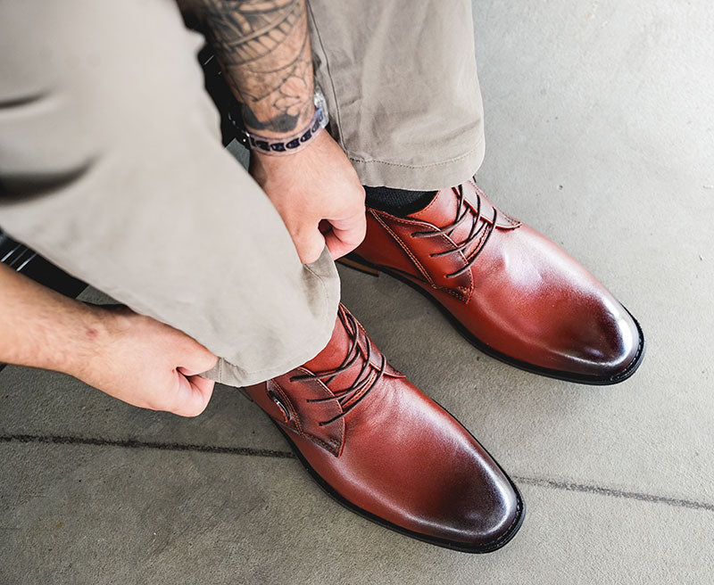 A close-up of a person tying brown leather dress shoes while seated, showcasing stylish footwear paired with beige pants.