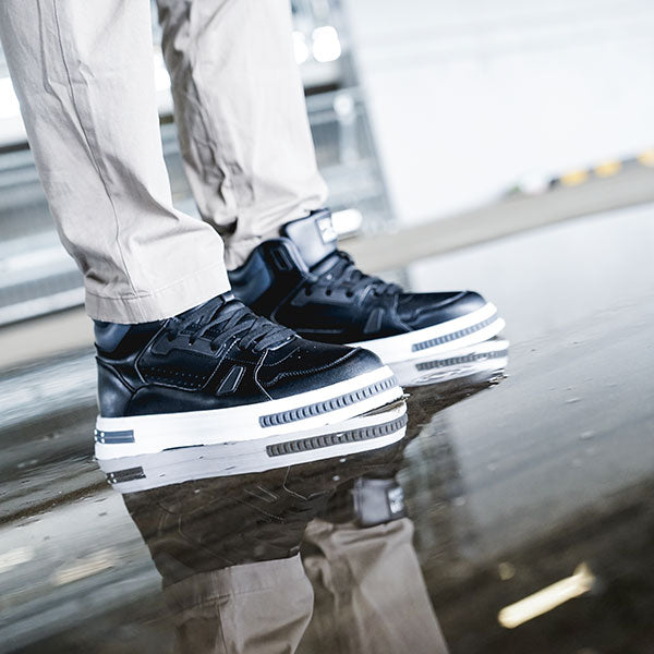 Close-up of stylish black sneakers with white soles, worn with light-colored pants, reflecting on a smooth floor.