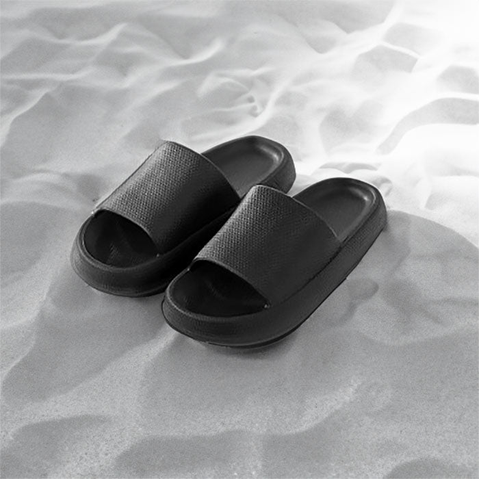A pair of black slide sandals with a textured top, resting on a sandy surface, ideal for casual summer wear.