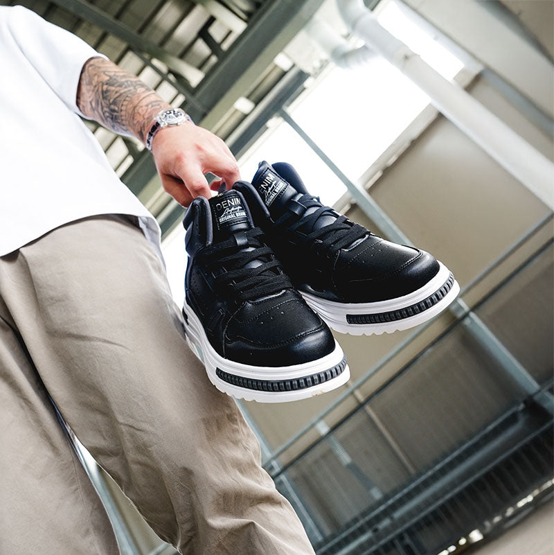 A person holds a pair of stylish black sneakers with white soles, showcasing their contemporary design against an industrial background.