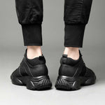 Black elevator sneakers worn with black pants on a neutral background