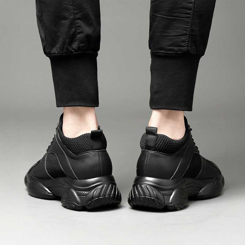 Black elevator sneakers worn with black pants on a neutral background