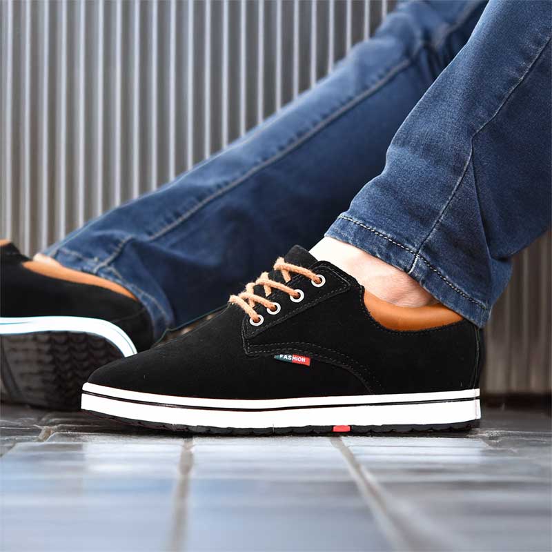 Black sneakers with white soles worn with blue jeans on a tiled floor.