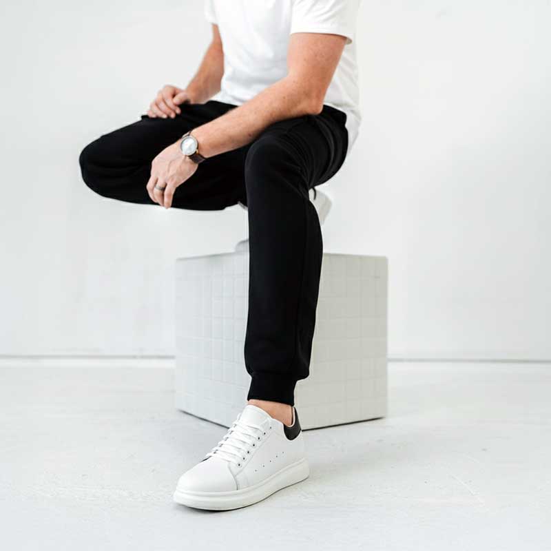 Person wearing black pants and white sneakers sitting on a white stool against a white background