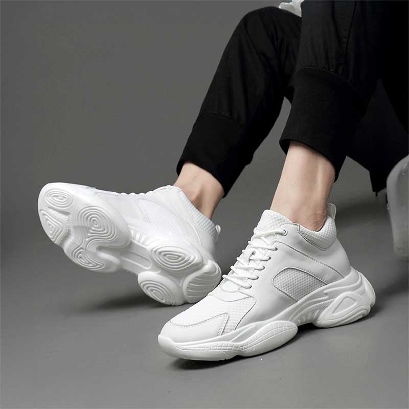 Man sitting in white elevator sneakers worn with black pants on a grey background
