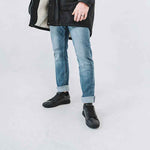A man in a black coat and rolled-up blue jeans stands on a white background, wearing stylish black sneakers.