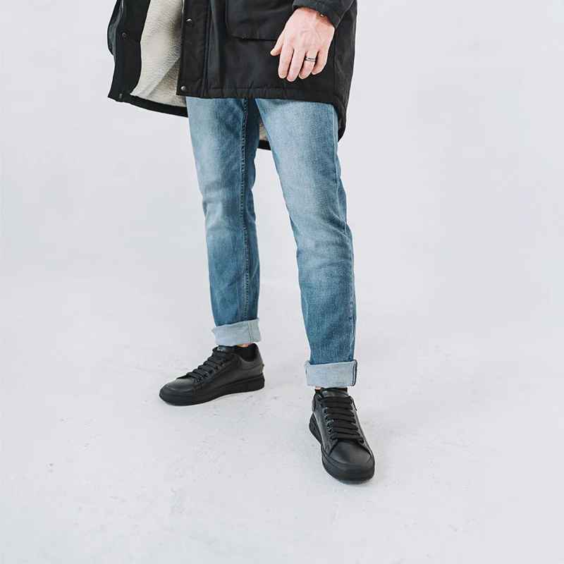 A man in a black coat and rolled-up blue jeans stands on a white background, wearing stylish black sneakers.