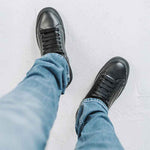 A pair of black sneakers worn with blue jeans, viewed from above on a light textured surface.